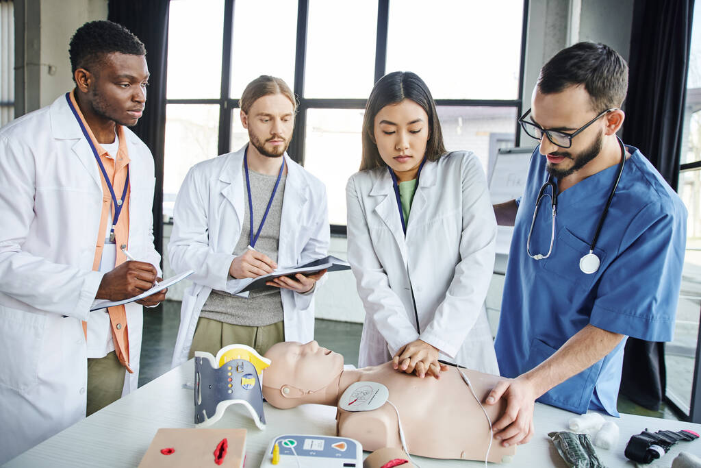 asian woman practicing chest compressions on CPR manikin near medical instructor and interracial students writing in notebooks during first aid seminar, emergency situations response concept - Photo, Image