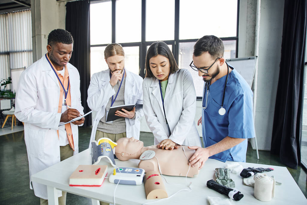 young asian student in white coat practicing chest compressions on CPR manikin near defibrillator, healthcare worker, and multicultural men with notebooks, emergency situations response concept - Photo, Image