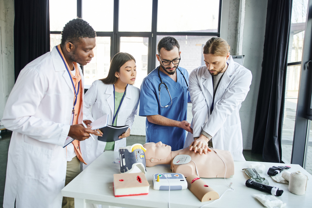 multiethnic students with notebooks looking at man doing chest compressions on CPR manikin near healthcare worker and medical equipment in training room, emergency situations response concept - Photo, Image