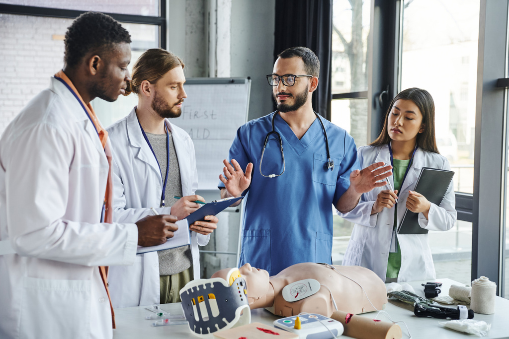 multicultural students in white coats writing near healthcare worker gesturing and talking next to CPR manikin and medical equipment on first aid seminar, emergency situations response concept - Photo, Image