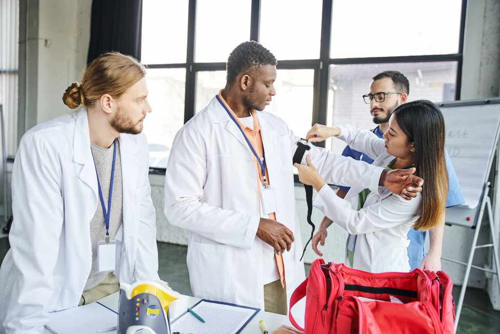 Ασιάτισσα με λευκό παλτό που εφαρμόζει αιμοστατικό επίδεσμο στο μπράτσο ενός Αφροαμερικανού φοιτητή κοντά σε εκπαιδευτή, ιατρικό εξοπλισμό και κουτί πρώτων βοηθειών, δεξιότητες διάσωσης και αντίληψη πρόληψης αιμορραγιών - Φωτογραφία, εικόνα