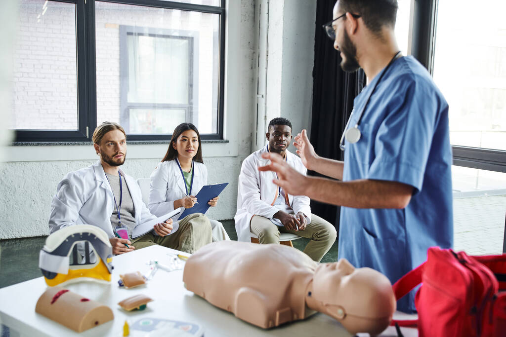 zdravotnický pracovník v modré uniformě hovořící s mnohonárodními studenty v bílých pláštích v blízkosti figuríny CPR a zdravotnického vybavení ve výcvikové místnosti, získávání a procvičování životně důležité dovednosti koncepce - Fotografie, Obrázek