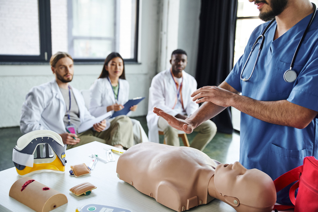 εργαζόμενος στον τομέα της υγειονομικής περίθαλψης που δείχνει τεχνικές διάσωσης στο ανδρείκελο CPR κοντά σε ιατρικό εξοπλισμό και ποικιλόμορφη ομάδα διαφυλετικών φοιτητών σε θολή βάση, αποκτώντας την έννοια των σωτήριων δεξιοτήτων - Φωτογραφία, εικόνα