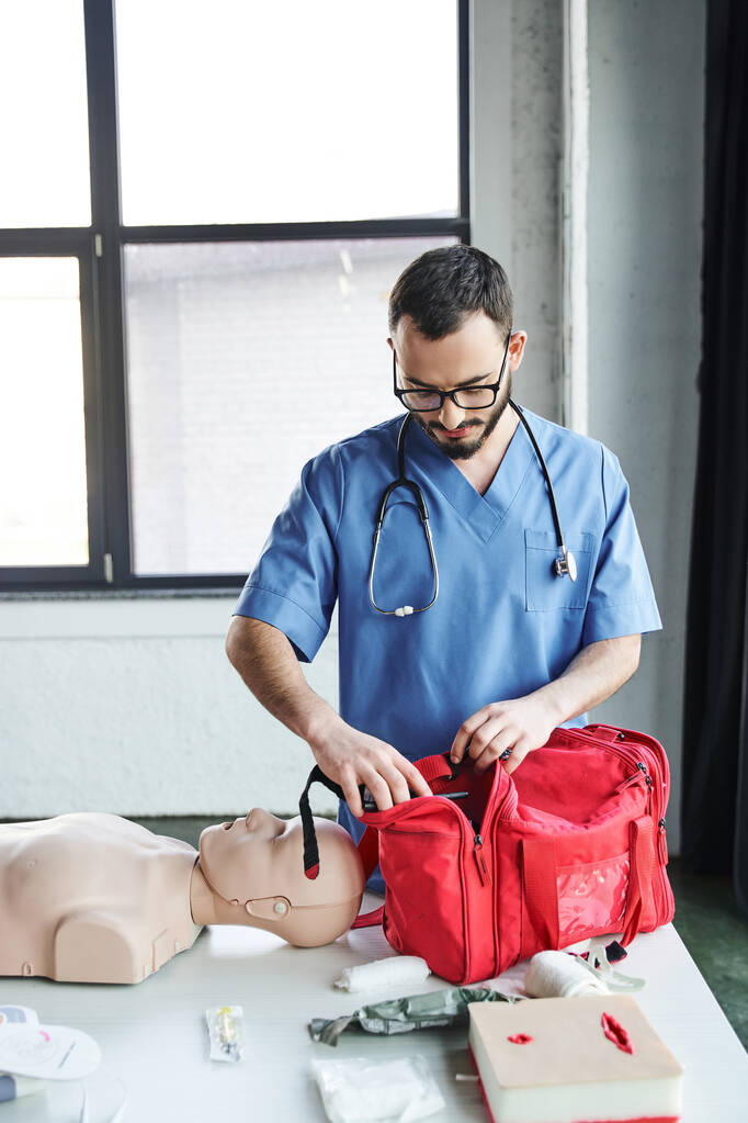 młody instruktor medycyny w okularach i niebieskim mundurze przygotowujący salę szkoleniową do seminarium pierwszej pomocy i otwierający czerwoną torbę koło manekina resuscytacyjnego, koncepcja rozwoju umiejętności ratujących życie - Zdjęcie, obraz