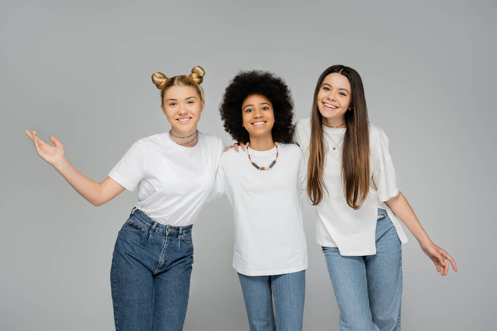 Cheerful and multiethnic teenage girlfriends in white t-shirts hugging each other and looking at camera while standing isolated on grey, teenage friends having fun together - Photo, Image