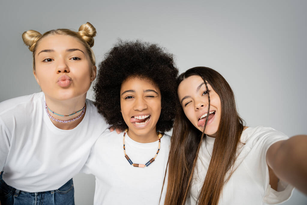 selfie, portret van vrolijke en multi-etnische tiener vriendinnen in witte t-shirts pruilen lippen en steken tongen terwijl knuffelen en poseren geïsoleerd op grijs, tiener vrienden samen plezier hebben - Foto, afbeelding