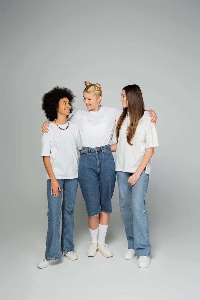 Full length of cheerful blonde teen girl hugging positive multiethnic girlfriends in white t-shirts and stylish blue jeans while standing on grey background, teenage friends having fun together - Photo, Image