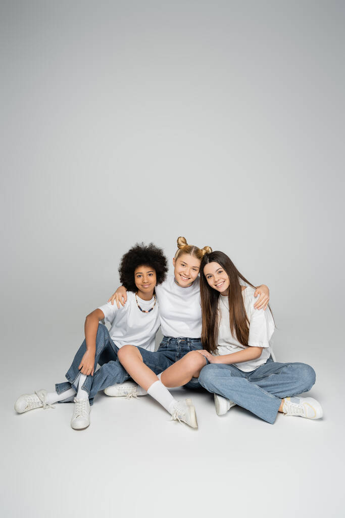 Πλήρες μήκος χαμογελαστές διαφυλετικές έφηβες φιλενάδες σε λευκά μπλουζάκια και μπλέ τζιν κοιτάζοντας την κάμερα ενώ κάθονται μαζί σε γκρι φόντο, έννοια πολυεθνικών εφηβικών μοντέλων - Φωτογραφία, εικόνα