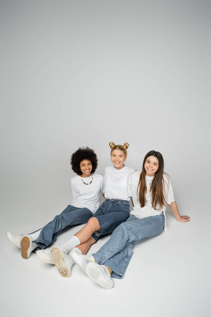 Veselé a multietnické dospívající přítelkyně v módních džínách a bílých tričkách při pohledu na kameru, zatímco sedí a pózují na šedém pozadí, multietnické teen modely koncept - Fotografie, Obrázek