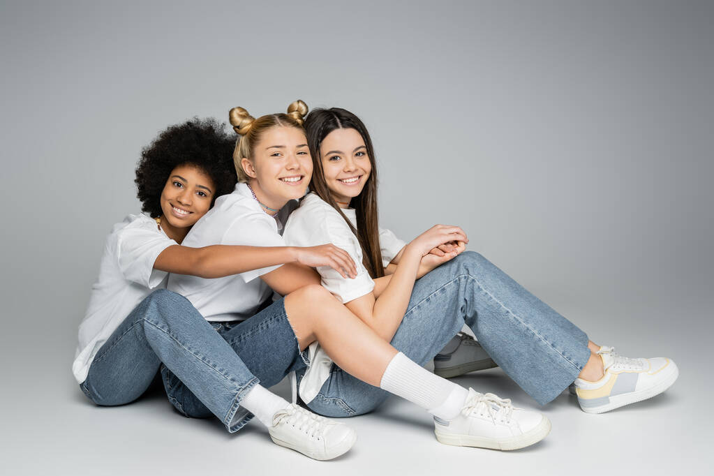 Полная длина положительных и многонациональных друзей-подростков в повседневных белых футболках и джинсах, смотрящих в камеру и сидящих на сером фоне, концепция многонациональных моделей подростков, дружба и связь - Фото, изображение