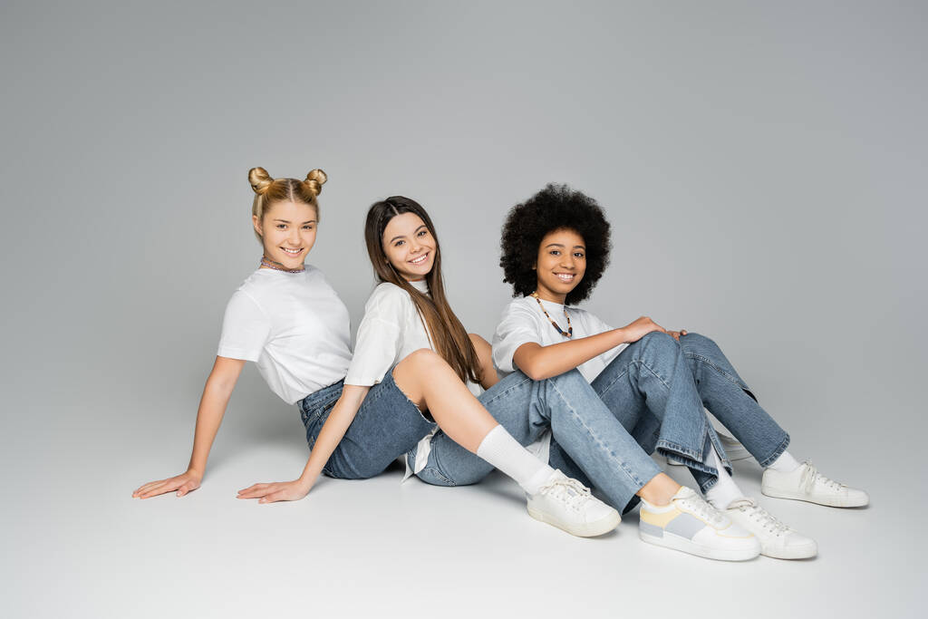 Volledige lengte van positieve en tiener multi-etnische vriendinnen in casual t-shirts en jeans kijken naar de camera terwijl samen zitten op een grijze achtergrond, multi-etnische tiener modellen concept - Foto, afbeelding