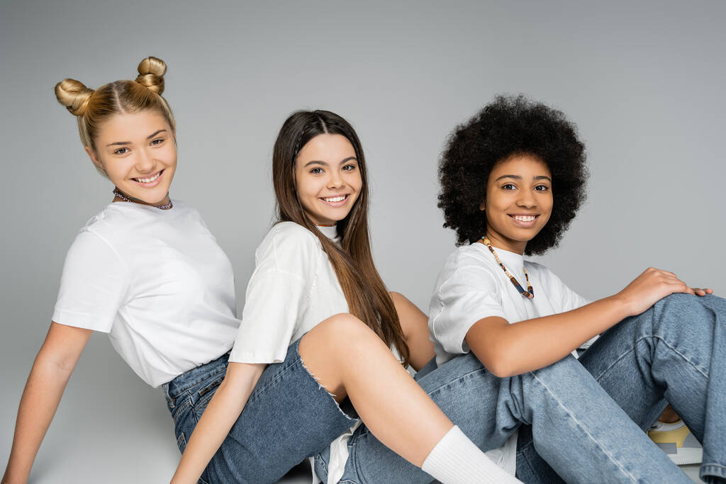 Χαρούμενες έφηβες πολυεθνικές φίλες με λευκά t-shirts και blue jeans που κοιτούν την κάμερα ενώ κάθονται και ποζάρουν μαζί σε γκρι φόντο, πολυεθνικά teen models concept, φιλία και δέσιμο - Φωτογραφία, εικόνα