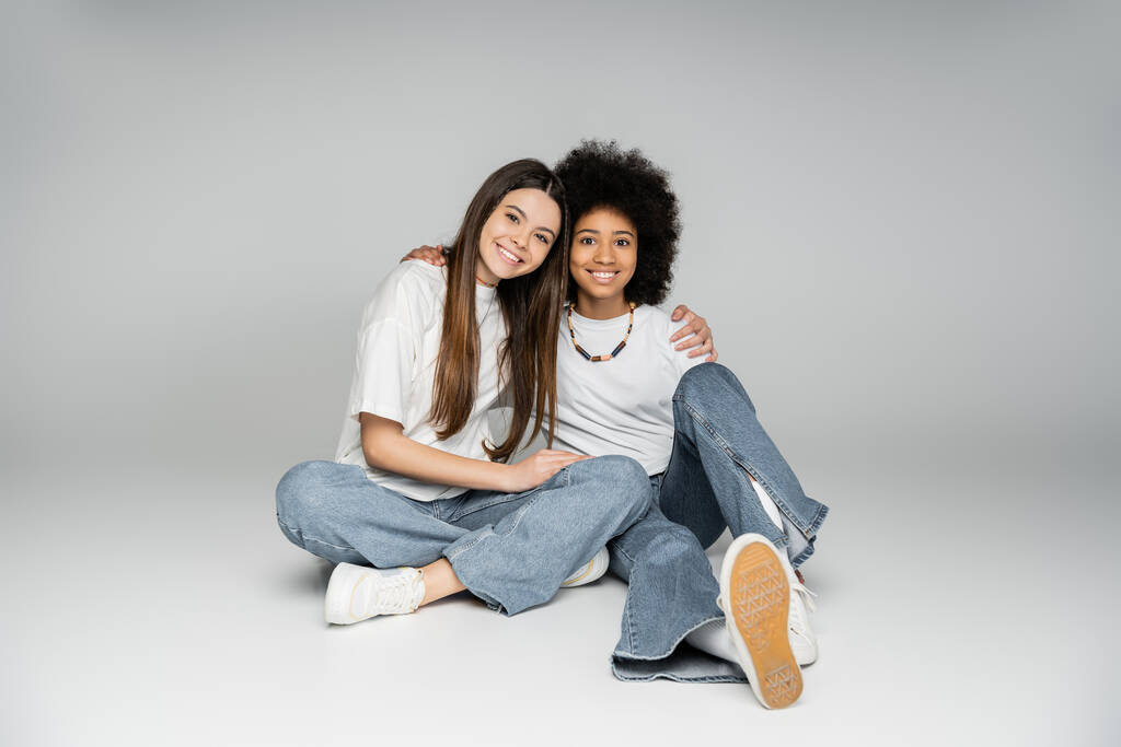 Lächelndes brünettes Teenie-Mädchen in weißem T-Shirt und Jeans umarmt afrikanisch-amerikanische Freundin, während sie zusammen sitzt und in die Kamera auf grauem Hintergrund schaut, lebhaftes Teenager-Mädchen-Konzept - Foto, Bild