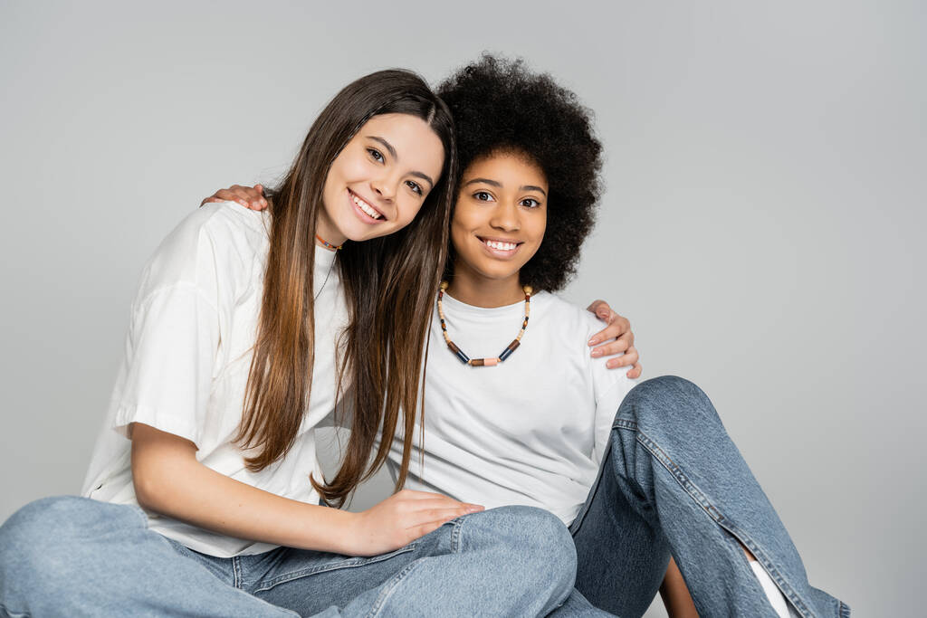 Joyful teen girl in casual white t-shirt and jeans hugging african american girlfriend and looking at camera while posing isolated on grey, lively teenage girls concept, friendship and bonding - Photo, Image
