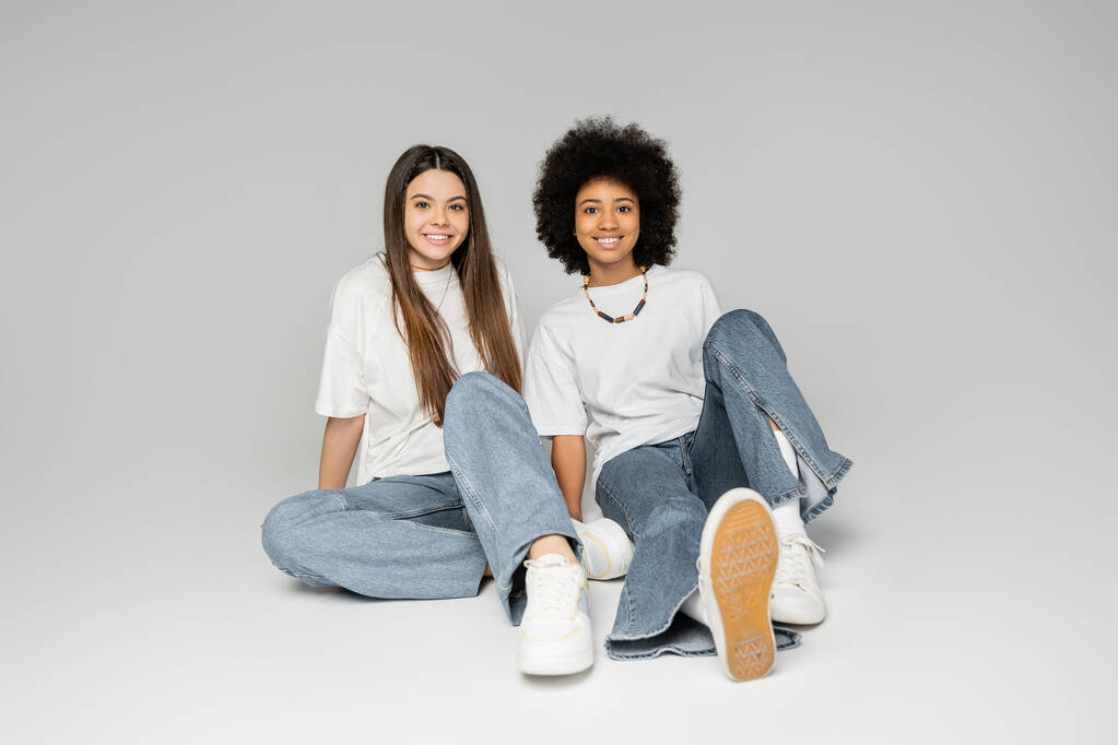 Full length of positive african american teenage girl in white t-shirt and blue jeans sitting next to brunette girlfriend on grey background, lively teenage girls concept, friendship and bonding - Photo, Image