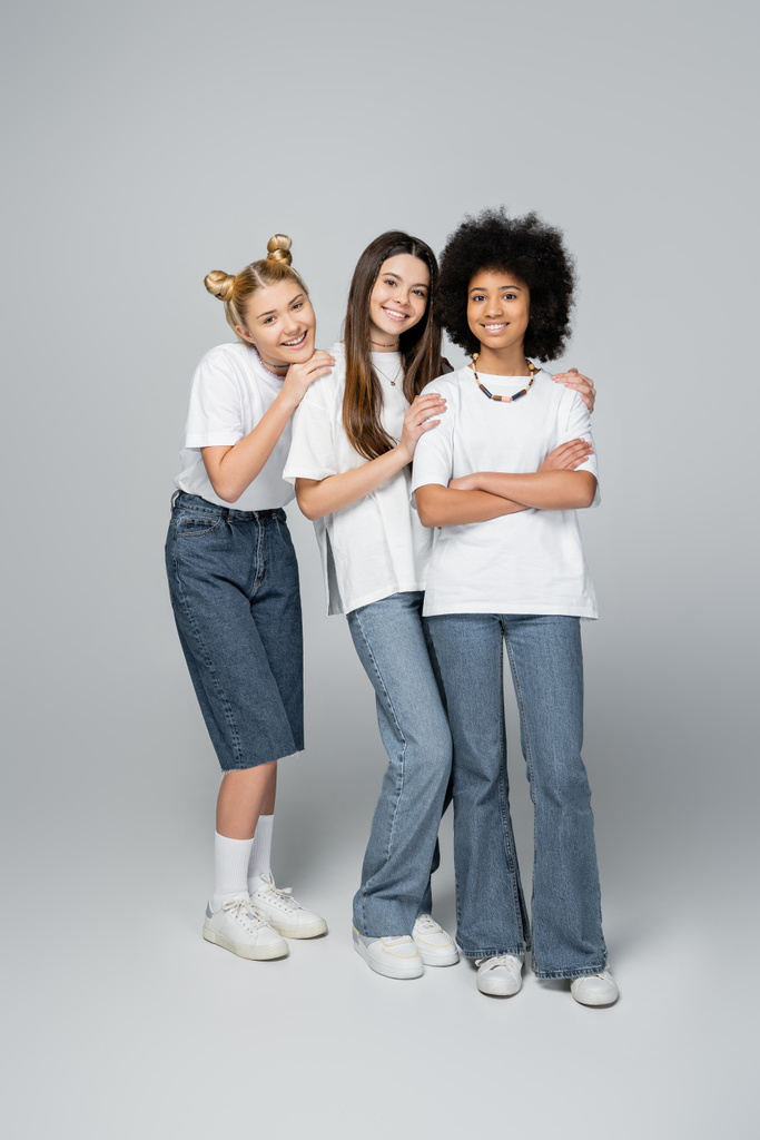Full length of positive and interracial teenagers in casual white t-shirts and jeans looking at camera and standing on grey background, lively teenage girls concept, friendship and bonding - Photo, Image