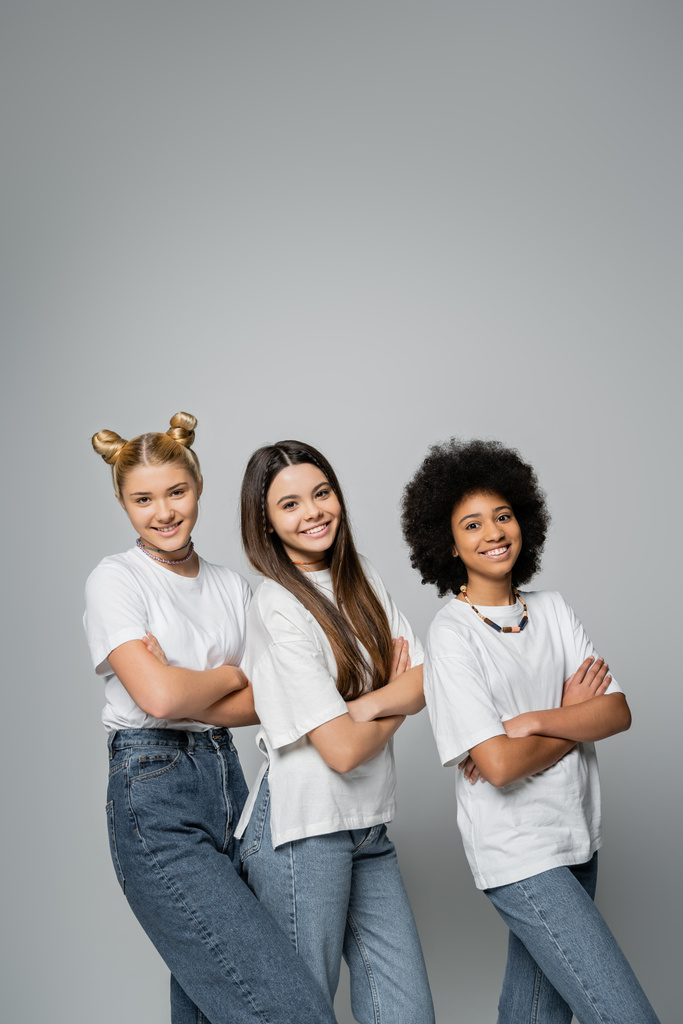 Positive and multiethnic teen girlfriends in stylish white t-shirts and jeans looking at camera and crossing arms while standing together isolated on grey, lively teenage girls concept - Photo, Image