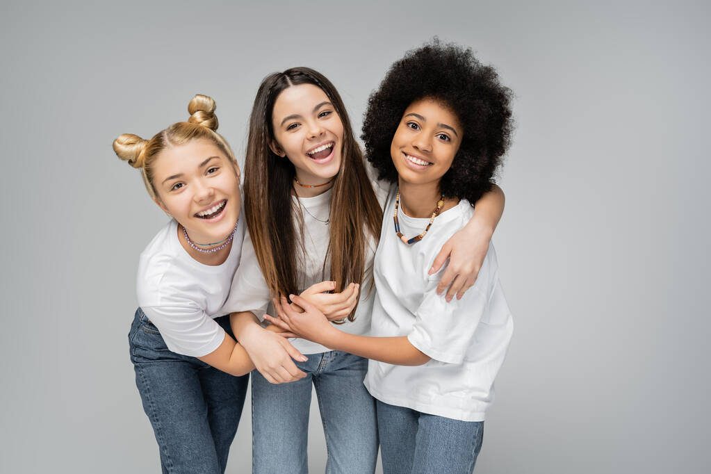 Positieve multi-etnische tiener meisjes in witte t-shirts en jeans knuffelen brunette vriend en kijken naar de camera terwijl staande geïsoleerd op grijze, levendige tienermeisjes concept, vriendschap en hechting - Foto, afbeelding