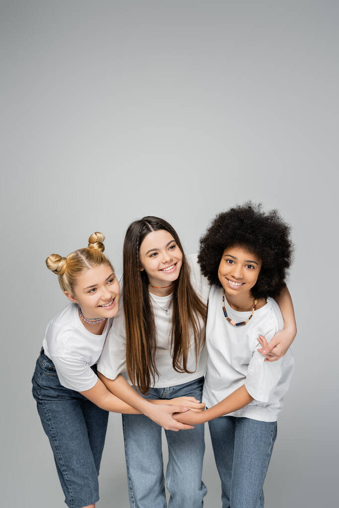 Namoradas adolescentes alegres e multiétnicas em camisetas brancas e jeans abraçando a amiga morena juntas e de pé isoladas no conceito, amizade e vínculo de meninas adolescentes cinzas e vivas - Foto, Imagem