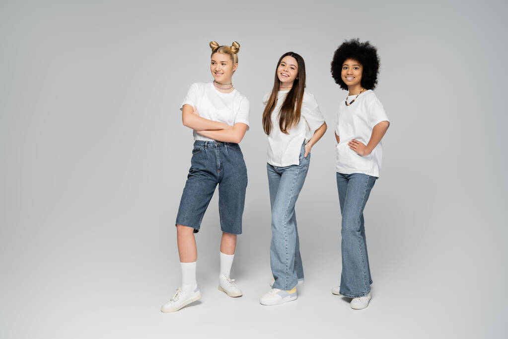 Volledige lengte van glimlachende interraciale tienermeisjes in witte t-shirts en jeans poseren en naar de camera kijken terwijl ze op een grijze achtergrond staan, levendig tienermeisjes concept, vriendschap en gezelschap - Foto, afbeelding