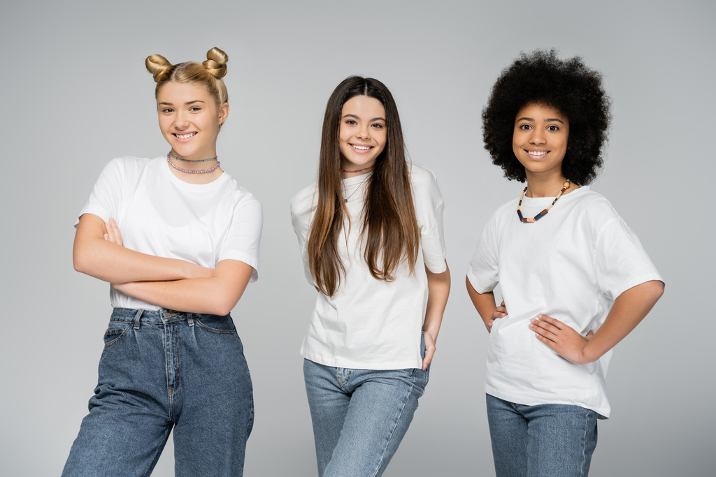 Positive and multiethnic teenage girls in jeans and casual white t-shirts posing and smiling at camera while standing isolated on grey, lively teenage girls concept, friendship and companionship - Photo, Image