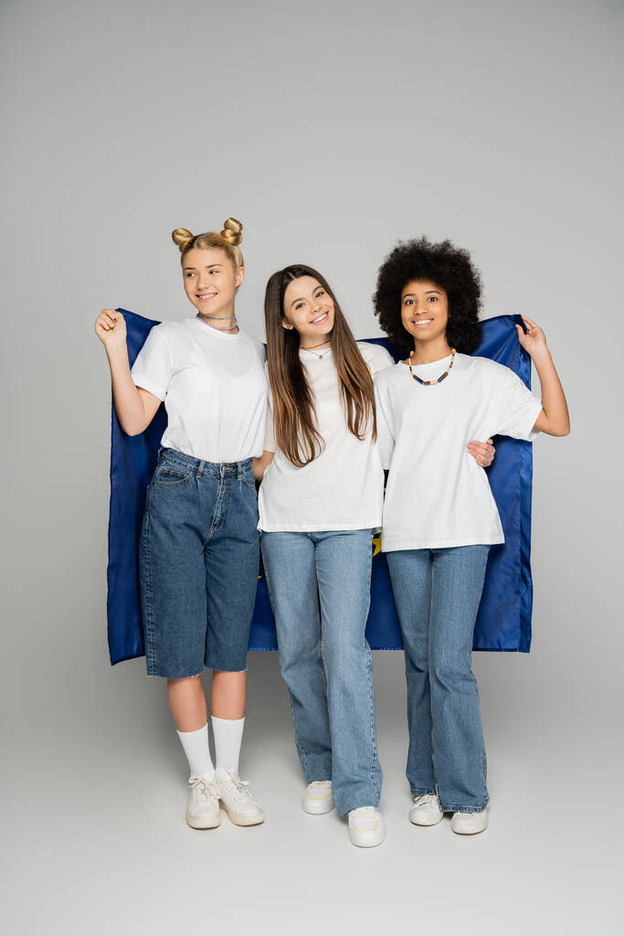 Pleine longueur de copines adolescentes souriantes et multiethniques en jeans et t-shirts blancs tenant le drapeau bleu et debout sur fond gris, des amis adolescents énergiques passent du temps - Photo, image