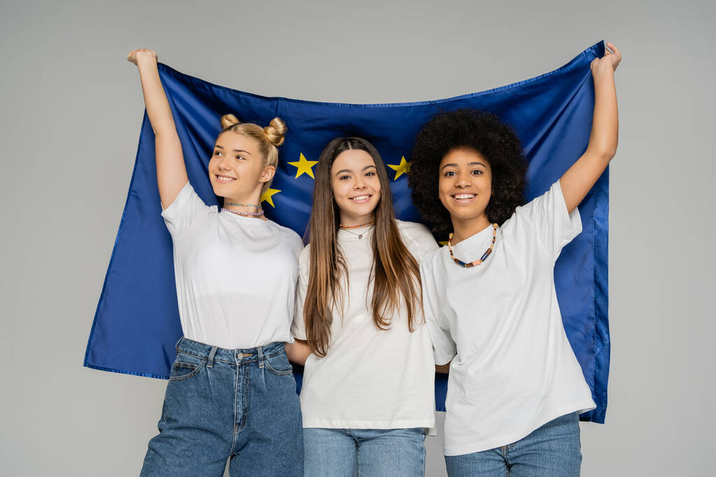 Pozitivní a multietnické dospívající dívky v bílých tričkách a džínách držící modrou evropskou vlajku, zatímco spolu stojí izolované na šedých, energických teenagerských přátelích trávících čas - Fotografie, Obrázek