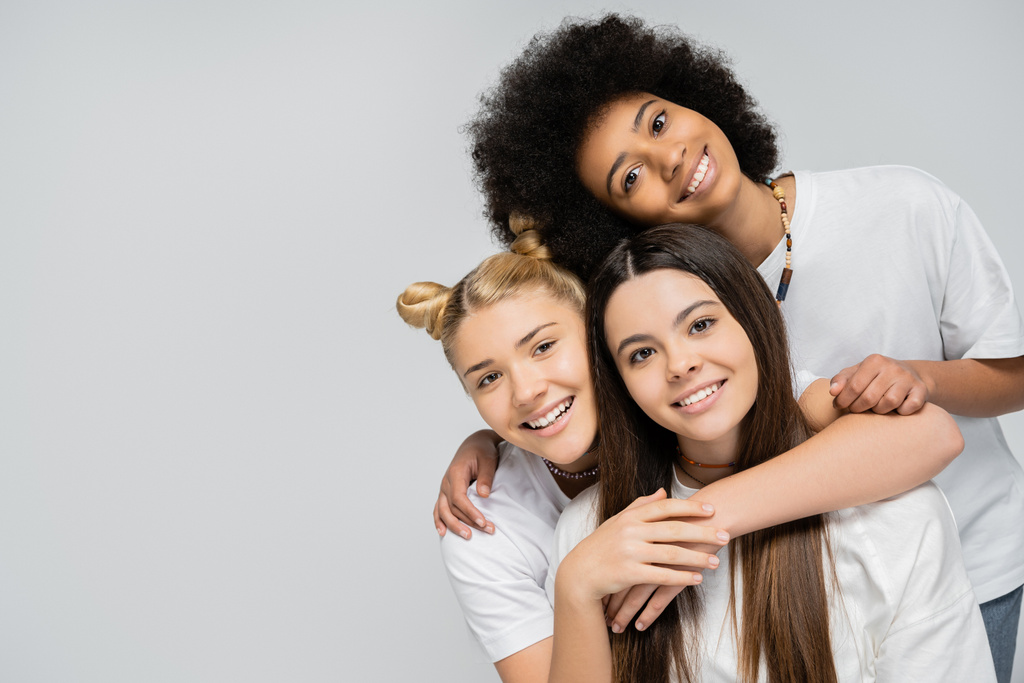 Gleeful and teenage girls in white t-shirts hugging each other and looking at camera while standing isolated on grey, energetic teenage friends spending time, copy space, friendship and companionship - Photo, Image