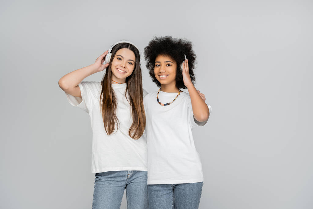 Joyful and interracial teen girls in white t-shirts and jeans listening music in headphones and looking at camera isolated on grey, energetic teenage models spending time, friendship and companionship - Photo, Image