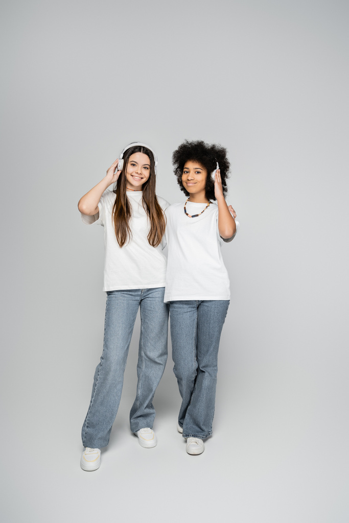 Pleine longueur de copines multiethniques et adolescentes en t-shirts et jeans blancs écoutant de la musique dans des écouteurs et debout sur fond gris, des modèles adolescents énergiques passent du temps  - Photo, image