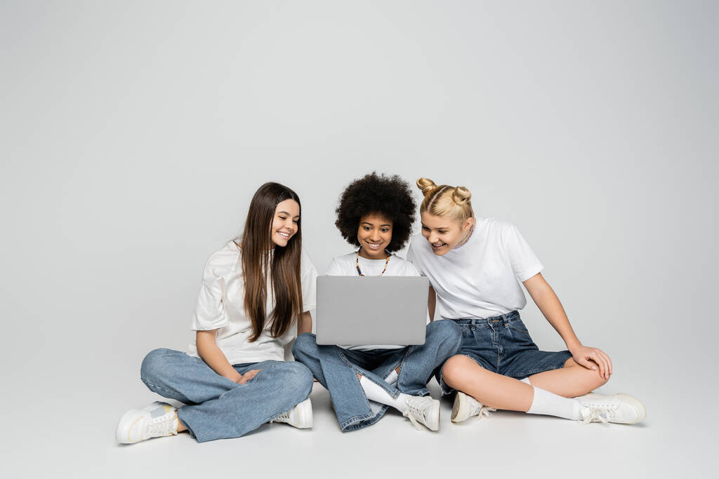 novias multiétnicas positivas en camisetas blancas y pantalones vaqueros usando el ordenador portátil juntos mientras están sentados en un fondo gris, los adolescentes se unen por el interés común, la amistad y la compañía - Foto, imagen