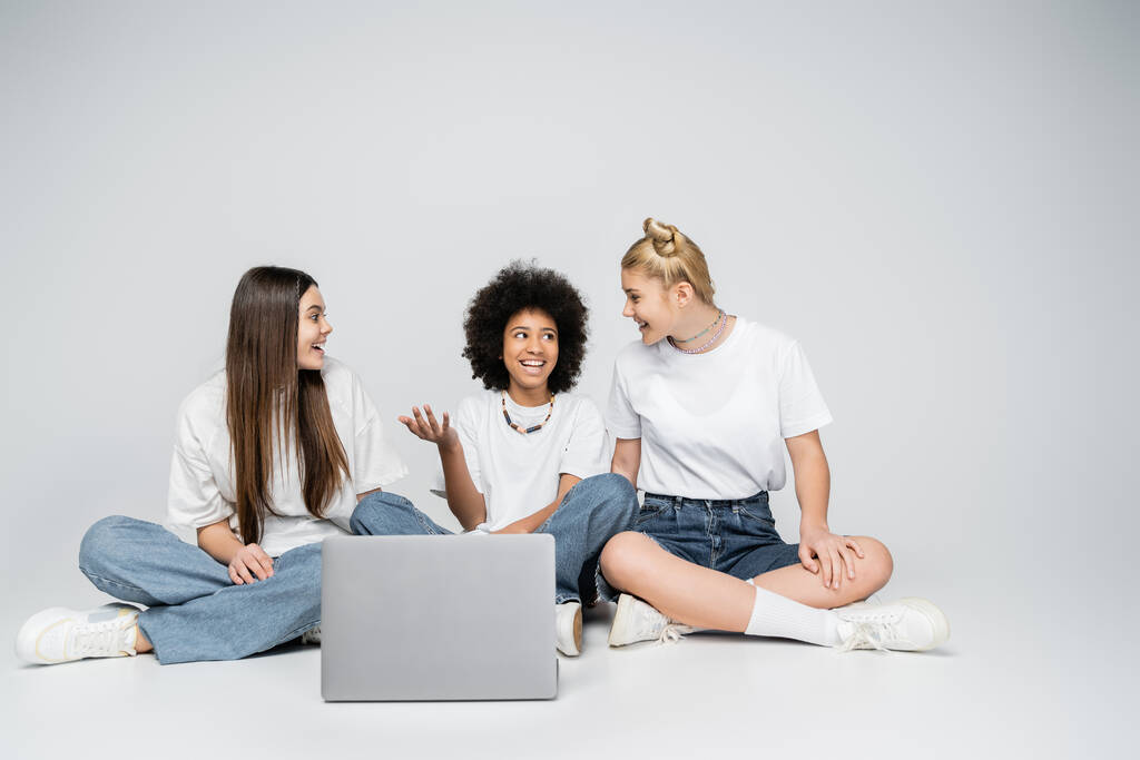 Wesoła afrykańska dziewczyna w białym t-shircie i dżinsach rozmawiająca z nastoletnimi dziewczynami siedząc razem przy laptopie na szarym tle, łącząca młodzież ponad wspólnym zainteresowaniem - Zdjęcie, obraz