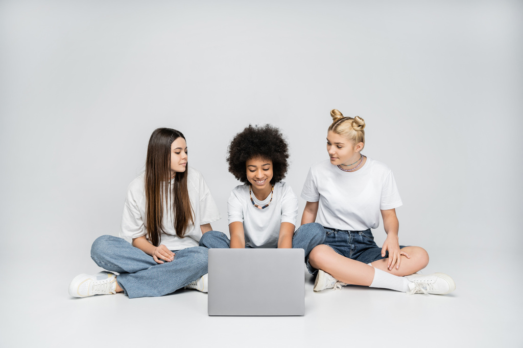 Smiling teen african american girl using laptop near girlfriends in white t-shirts and jeans and sitting together on grey background, teenagers bonding over common interest - Photo, Image