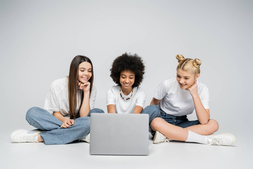 Teen african american girl using laptop near girlfriends in white t-shirts and jeans while sitting together on grey background, teenagers bonding over common interest, friendship and companionship - Photo, Image