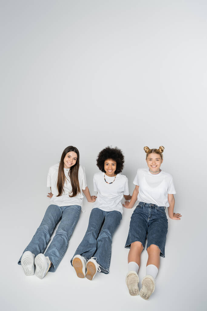 Vue grand angle de copines adolescentes souriantes et multiethniques en t-shirt blanc et jeans regardant la caméra tout en étant assis sur fond gris, modèles d'adolescence et concept de génération z - Photo, image