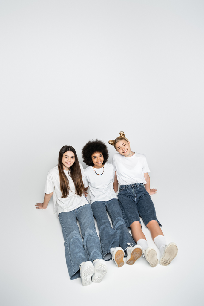 白いTシャツとジーンズを着た正と多民族の十代の友人の高い角度のビューは、カメラを見て、灰色の背景、思春期のモデルと世代のzの概念に一緒に座って - 写真・画像