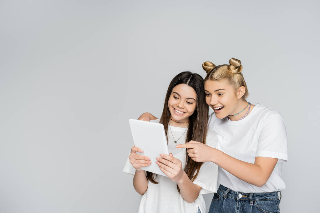 Usmívající se blondýnka teen dívka v džínách a bílém tričku ukazuje prstem na digitální tablet a objímající brunetka přítel izolované na šedé, teenageři sbližování nad společným zájmem, Touch Screen - Fotografie, Obrázek