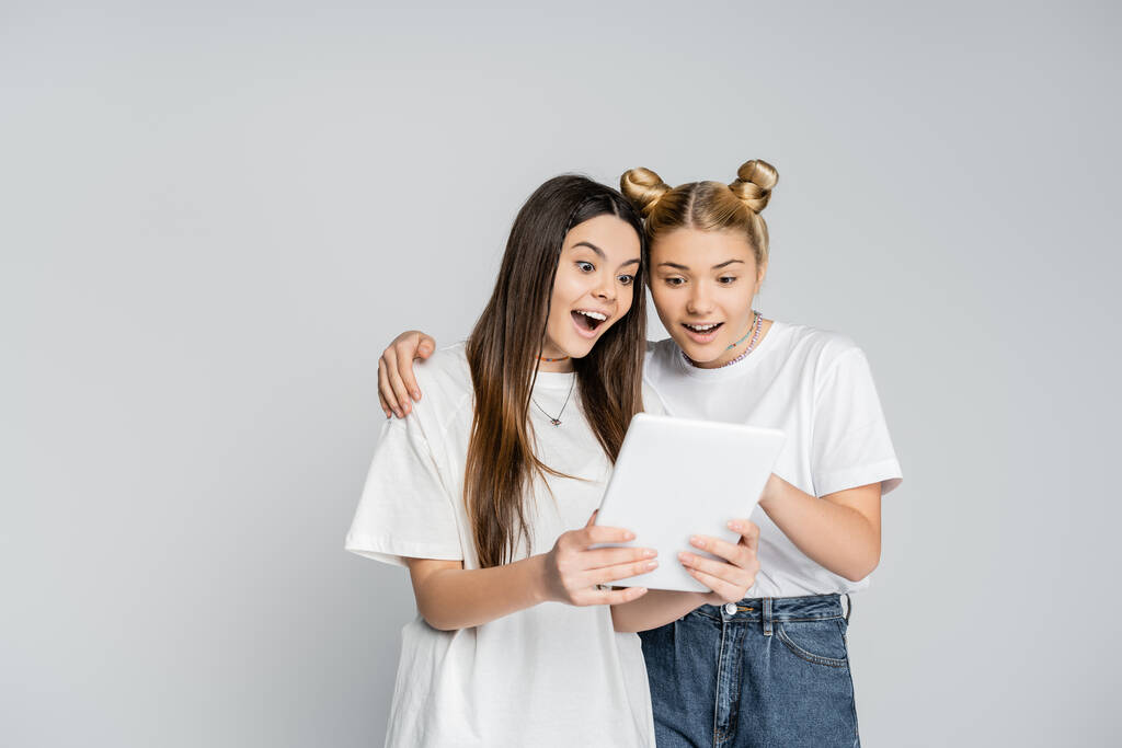 Aufgeregte Teenager-Freundinnen in weißen T-Shirts, die gemeinsam ein digitales Tablet benutzen, während sie isoliert auf grauen Stühlen stehen, Teenager, die sich über gemeinsame Interessen, Freundschaft und Kameradschaft einig sind. - Foto, Bild