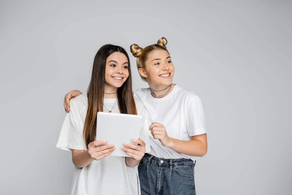 Усміхнена блондинка дівчина-підліток у білій футболці і джинсах обіймає брюнетку подругу з цифровим планшетом і дивиться разом ізольовано на сірому, підлітків, що зв'язуються над загальними інтересами
 - Фото, зображення