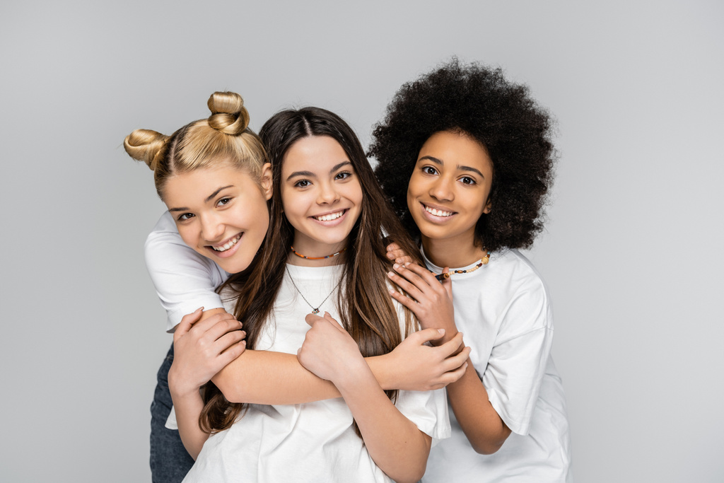 Portret pozytywnych i międzyrasowych nastolatek w białych t-shirtach przytulających brunetkę i patrzących na odizolowaną kamerę na szarych modelkach z okresu dojrzewania i koncepcji generacji z - Zdjęcie, obraz
