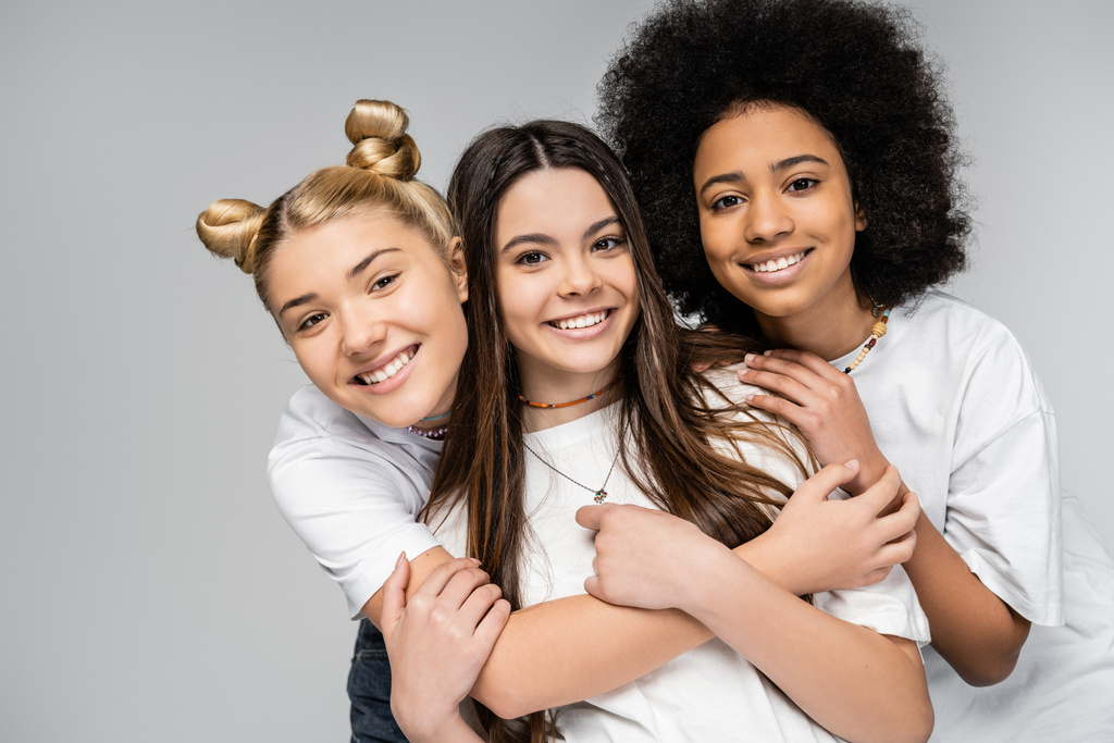 Portrait d'adolescentes gaies et multiethniques en t-shirts blancs s'embrassant et regardant la caméra tout en étant isolées sur des amis gris et adolescents s'amusant ensemble - Photo, image