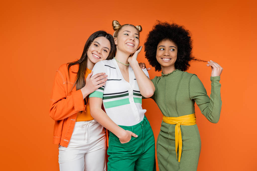 Usmívající se multietnické teen dívky s tučným make-up pózovat v blízkosti trendy blondýny přítelkyně a stojí spolu na oranžovém pozadí, kulturní rozmanitost a generace z módní koncept - Fotografie, Obrázek