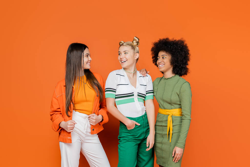 Wesołe i wieloetniczne dziewczyny w modnych strojach rozmawiające i patrzące na siebie stojąc razem na pomarańczowym tle, różnorodności kulturowej i koncepcji mody pokolenia z - Zdjęcie, obraz