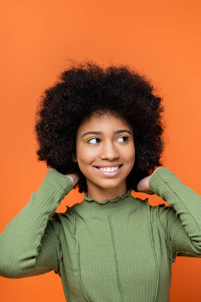 Portrait of joyful teenage african american girl with bold makeup and green dress touching hair and looking away isolated on orange, teenage fashion and generation z concept  - Photo, Image