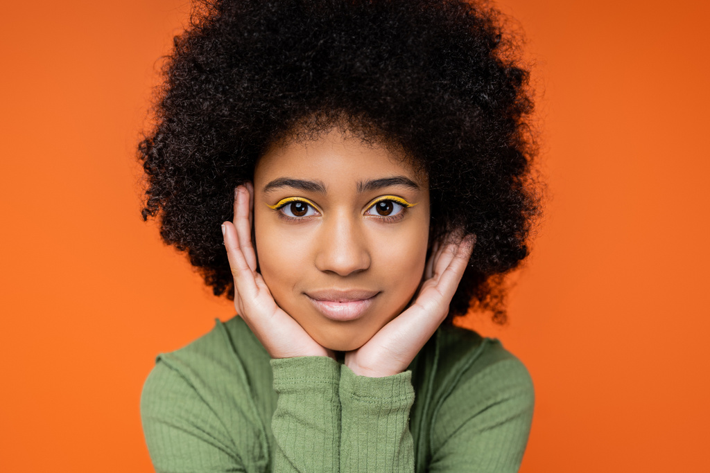 Portrait of teenage african american girl with bright makeup wearing green dress and looking at camera while touching cheeks isolated on orange, youth culture and generation z concept  - Photo, Image