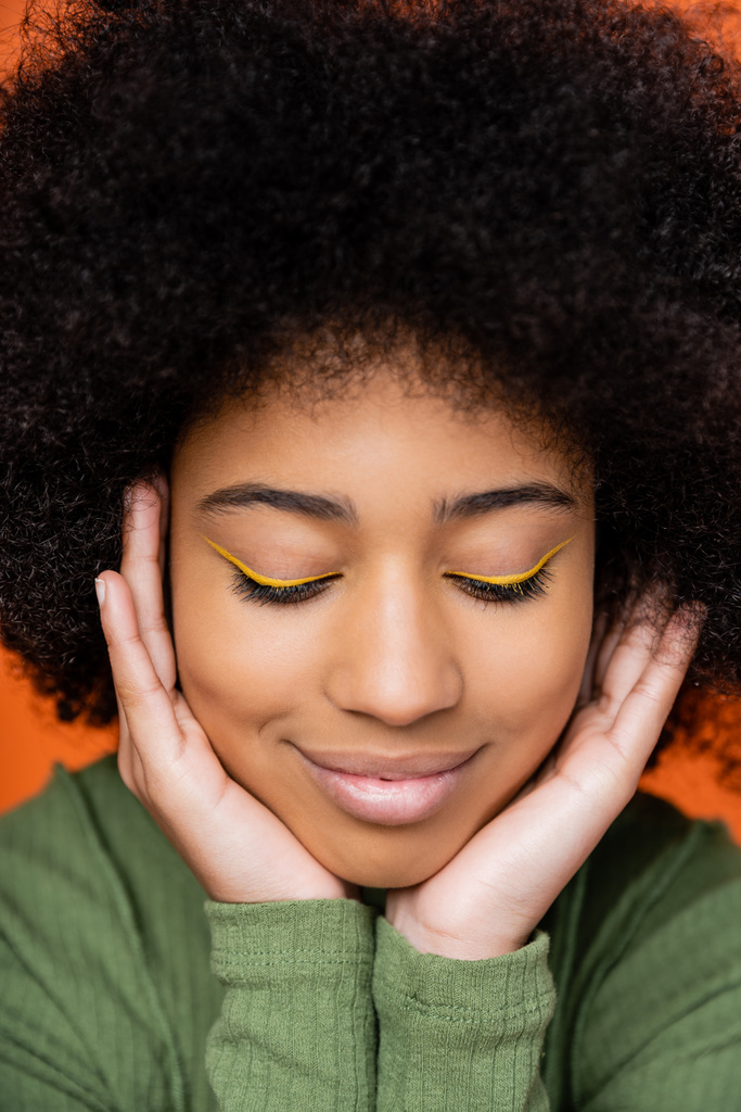 Retrato de sorrir e adolescente na moda menina americana africana com maquiagem brilhante tocando bochechas e posando isolado em laranja, cultura juvenil e conceito z geração  - Foto, Imagem