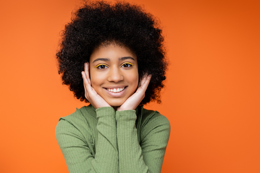 Portret szczęśliwej nastolatki amerykańskiej dziewczyny z odważnym makijażem ubranym w zieloną sukienkę i dotykającymi policzkami stojąc wyizolowany na pomarańczowej, młodzieżowej kulturze i koncepcji pokolenia z  - Zdjęcie, obraz