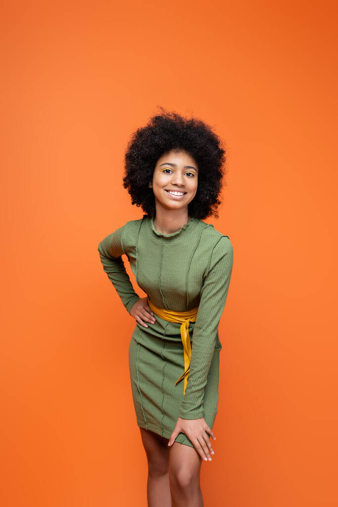 Chica afroamericana adolescente moderna y positiva con maquillaje brillante usando vestido verde y sosteniendo la mano en la cadera aislada en naranja, cultura juvenil y concepto de generación z  - Foto, Imagen