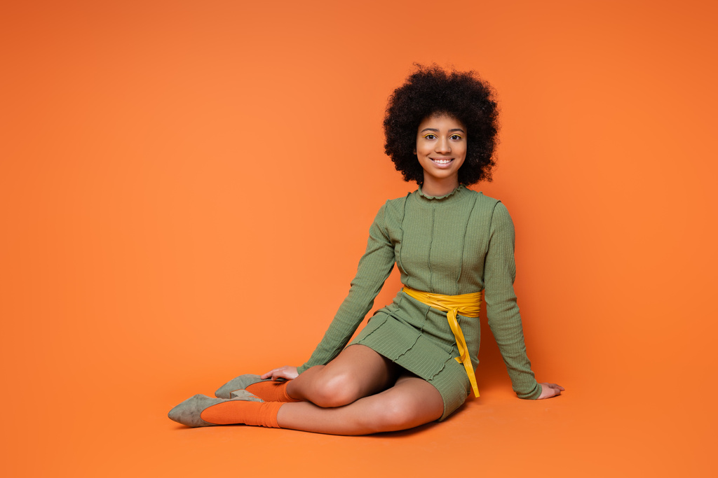 Cheerful and teen african american girl in fashionable green dress smiling at camera while sitting and posing on orange background, youth culture and generation z concept  - Photo, Image