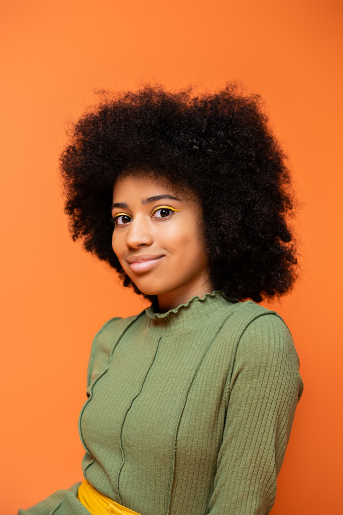 Porträt eines modischen afrikanisch-amerikanischen Teenie-Mädchens mit kühnem Make-up in grünem Kleid und lächelnd in die Kamera isoliert auf orange, Jugendkultur und Generation-Z-Konzept  - Foto, Bild